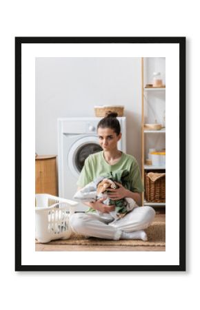 puzzled young woman holding clothes near basket and washing machine in laundry room.