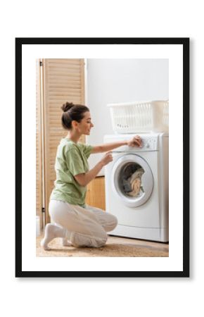 Side view of smiling woman switching washing machine near basket at home.