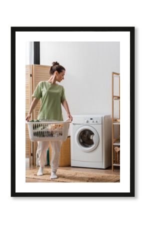 Side view of young woman holding basket with clothes and looking at washing machine at home.