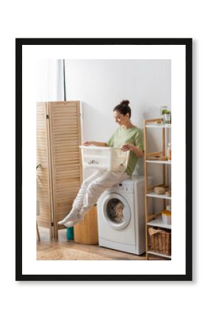 Young woman looking at basket while sitting on washing machine in laundry room.