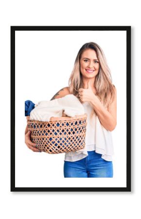 Young beautiful blonde woman holding laundry basket smiling happy and positive, thumb up doing excellent and approval sign