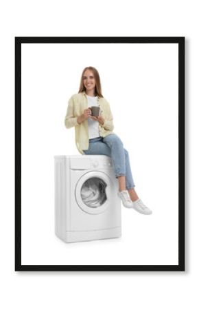 Beautiful young woman with cup of drink on washing machine with laundry against white background