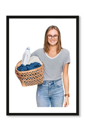 Beautiful blonde woman holding laundry basket and detergent bottle looking positive and happy standing and smiling with a confident smile showing teeth