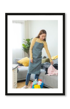 Household clean up, housekeeper asian young woman cleaning, hand keeping put clothes in basket on sofa in living room at home. Mess maid or housewife organizing dirty and untidy. Chore concept.
