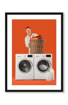 Handsome young man holding basket with laundry near washing machines on orange background