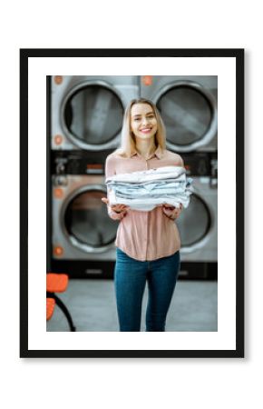 Portrait of a young woman holding ironed clothes in the professional laundry with drying machines on the background