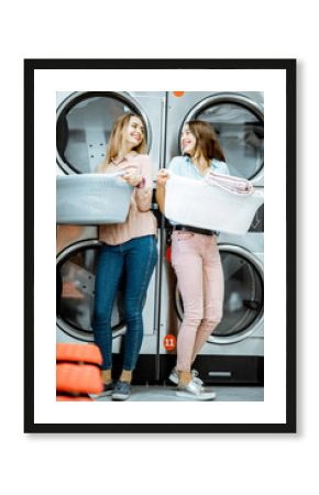 Two young women standing together with baskets full of clothes in the laundry with dryer machines on the background