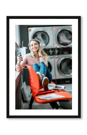 Young woman enjoying music waiting for the clothes to be washed sitting on the chair at the self-service laundry