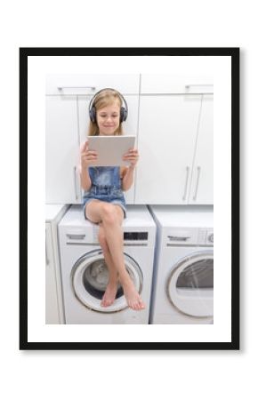 a Happy young girl listens to music on headphones holding tablet in laundry room with washing machine