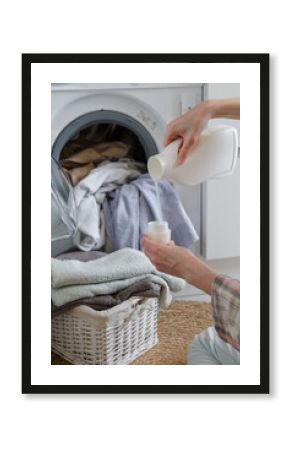 Close up of female hands pouring liquid laundry detergent into cap. Washer machine and clothes with wicker basket in background