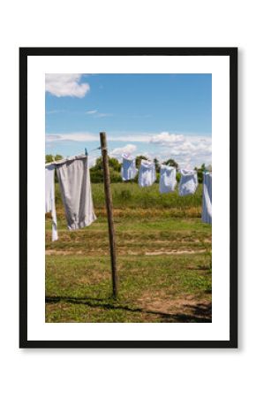 White laundry hangs on a clothesline strung between wooden poles in a sunny rural setting in Italy, with a blue sky and scattered clouds above a green and brown field.