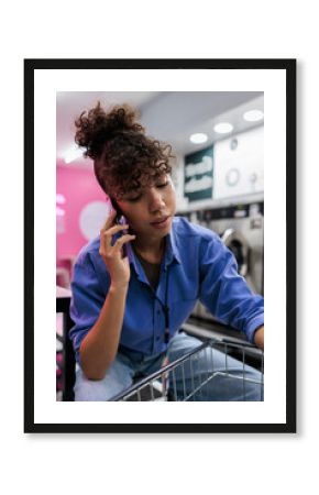 Woman on phone at laundromat, sitting on laundry cart.