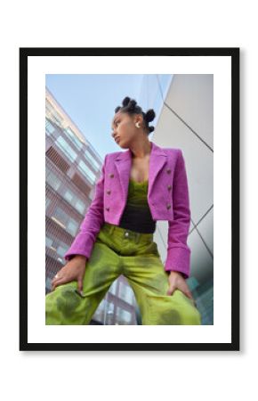 Vertical shot of stylish woman dressed in pink jacket and green trouers leans on knees focused away poses against urban buildings in city notices something. People fashion and lifestyle concept