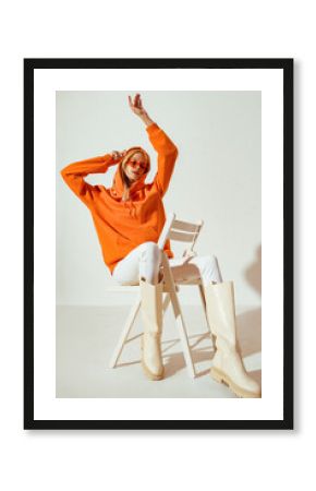 Full-length studio fashion portrait of playful confident woman wearing stylish orange hoodie, sunglasses, white skinny jeans, high boots, sitting, posing on chair. 