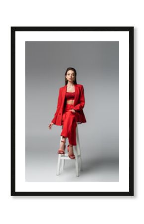 full length of pretty woman in red suit and shoes sitting on white chair on dark grey.