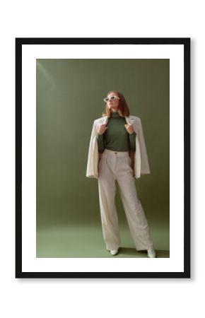 Fashionable confident woman wearing elegant white suit with blazer, wide leg trousers, cashmere turtleneck sweater, trendy sunglasses, posing on green background. Full-length studio fashion portrait