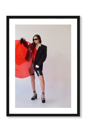 beautiful asian woman in stylish look and sunglasses posing holding red round shaped glass, grey background, blazer and latex shorts, youthful model, fashion forward, studio photography
