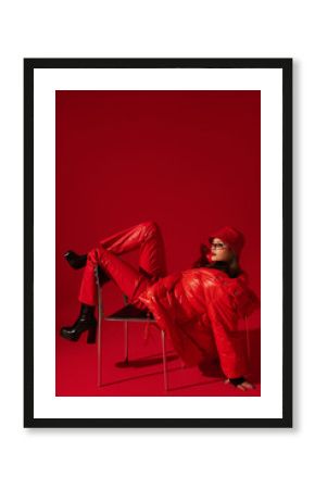 Fashionable confident woman wearing trendy oversized red puffer down jacket, sunglasses, bucket hat, faux leather pants, platform boots, posing on red background. Full-length studio fashion portrait