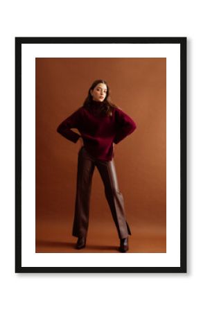 Fashionable confident woman wearing burgundy color knitted sweater, faux leather pants, pointed toe boots, posing on brown background. Full-length studio fashion portrait