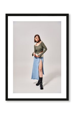 A young woman with brunette hair poses stylishly in a skirt and boots in a studio setting.