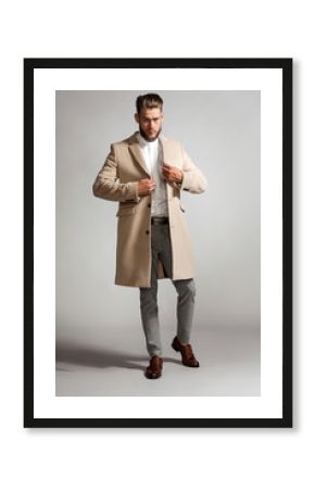 Elegant bearded man in a suit zips a beige coat, standing on a gray background