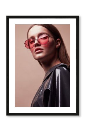 Fashion portrait of beautiful young woman in round red sunglasses and grey latex jacket posing in studio