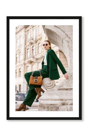 Outdoor full body fashion portrait of  fashionable woman wearing sunglasses, white turtleneck, green suit, blazer, trousers, leopard print boots, holding suede bag, posing in street of european city