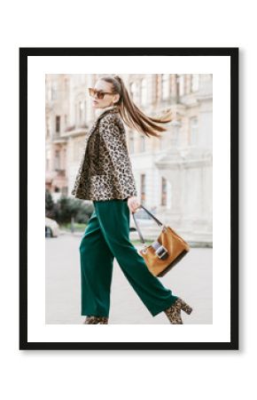 Outdoor full body fashion portrait of young beautiful fashionable woman wearing sunglasses, leopard print blazer, boots, green trousers, holding brown suede bag, walking in street of european city