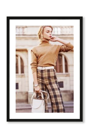 Outdoor fashion portrait of young beautiful fashionable girl wearing trendy beige cashmere turtleneck, high-waisted checkered trousers, belt, wrist watch, holding small white bag, posing in street