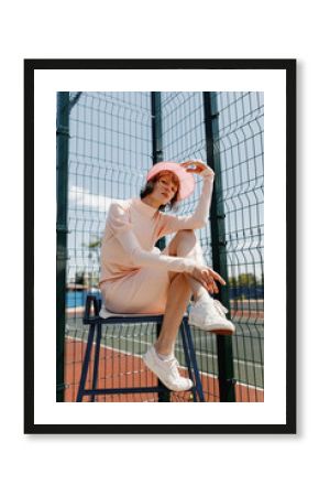 Stylish girl in a gently pink dress and pink visor sitting on a chair behind fence of the lattice at the sports field on a summer sunny day