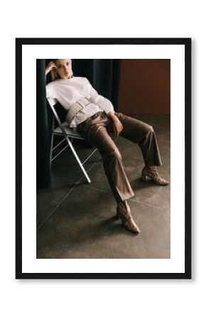 stylish blonde woman in white blouse and boots with snakeskin print sitting on chair near curtain on brown