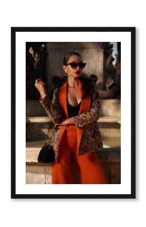 Fashion style outdoor portrait. Elegant woman in colorful orange suit with black handbag standing and posing against fountain in old town on a sunny evening, Sunny evening. Tropical city