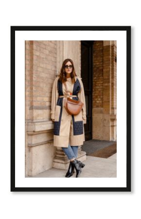 street style portrait of an attractive woman wearing a beige trench coat, denim jeans, black ankle boots and brown leather bag, crossing the street. fashion outfit perfect for autumn fall winter