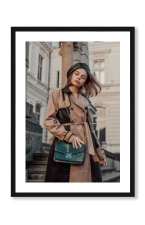 Outdoor autumn fashion portrait of young elegant confident woman wearing beige black trench coat, leather beret, holding green faux leather textured crocodile bag, posing in street of European city