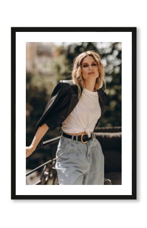 Stylish fashionable blonde woman with smoky eye makeup, in jeans, white t-shirt and black leather jacket on the balcony in the city. Spring autumn fashion concept. Soft selective focus.