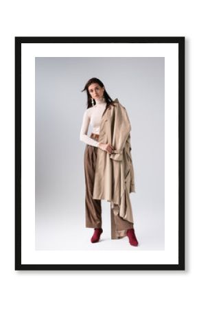 full length view of attractive, trendy girl with trench coat on shoulder looking at camera on grey