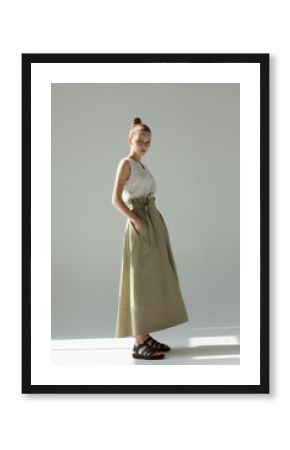 Young woman posing in studio on a light background. Long green skirt, white summer T-shirt, black sandals. She stands sideways, hands in pockets.