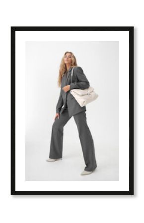 High fashion full length portrait of young beautiful female blonde model in classic gray male suit. Vogue style. Girl with blue eye arrows standing on white background and holding quilted handbag