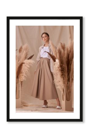 Young elegant female model standing on beige textile background and posing between ears of rye. Brunette girl with makeup and pony tail hair in white shirt and beige skirt and jacket