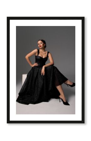 Elegant lady in black shiny ball gown and shoes sitting at white cibe with crossed legs and posing against grey background, Full length fashion portrait. Sexy female model