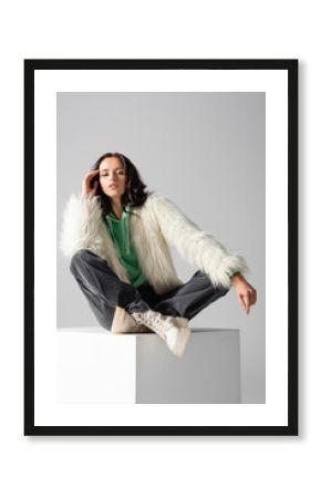  young woman in faux fur jacket posing on cube on white background