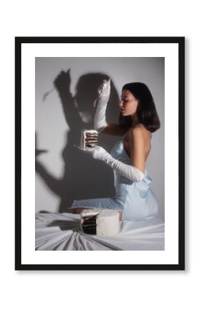 Stylish brunette in a silky blue dress and satin gloves poses with a slice of cake under dramatic shadows. A conceptual fashion and boudoir image ideal for editorial or promotional use.