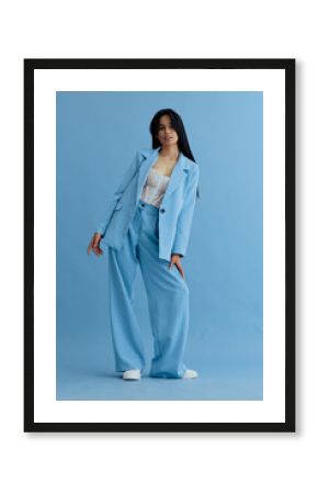 Young woman in stylish light blue suit posing confidently with casual lace top and white sneakers against a matching blue studio background, exuding modern fashion vibe.