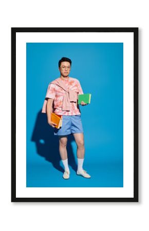 Stylish young Asian man showcasing relaxed fashion with colorful books against blue backdrop
