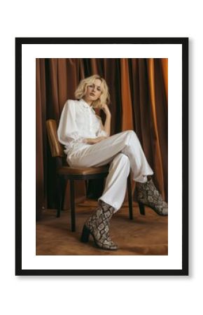 Blonde woman sits on brown chair near curtain. White blouse, boots with snakeskin print, stylish outfit. Young adult, caucasian, fashion model, posing, copy space.