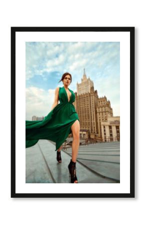 Attractive brunette girl in a long emerald dress posing on the catwalk on the roof against the background of the city, fashion concept, model in a luxurious dress against the background of urban