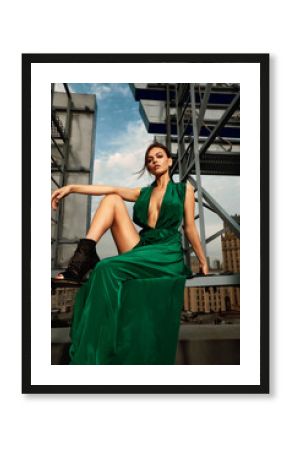 Attractive brunette girl in a long emerald dress posing on the catwalk on the roof against the background of the city, fashion concept, model in a luxurious dress against the background of urban