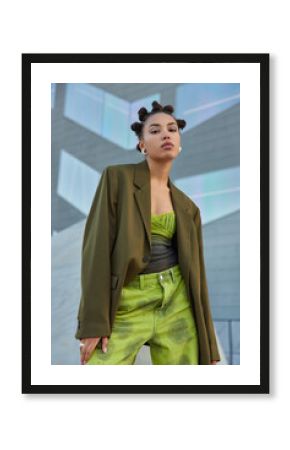 Self confident fashionable teenage girl dressed in green jacket and jeans has funny hairstyle poses against grey wall in public place has serious expression. Youth fashion and style concept.