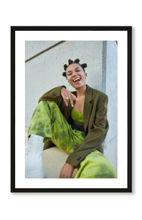 Vertical shot of overjoyed carefree teenage girl wears stylish green clothes laughs happily poses against concrete wall feels free spends free time outdoors. People fashion and style concept