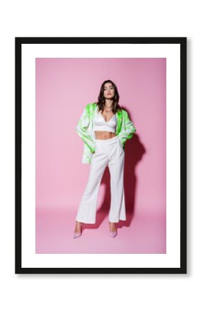 full length of brunette woman in stylish outfit posing with hands on hips on pink